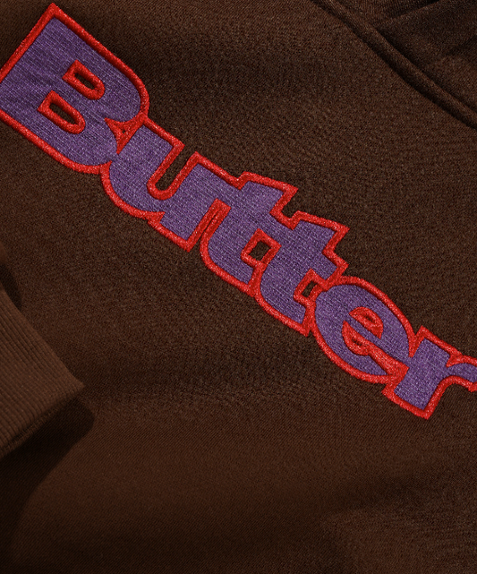 Close-up of a brown fleece or sweatshirt fabric with a large embroidered 'Butter' logo in purple with a red outline.