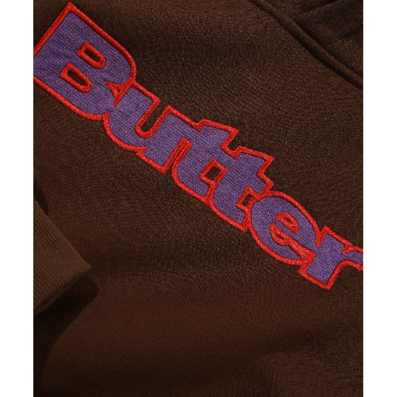 Close-up of a brown fleece or sweatshirt fabric with a large embroidered 'Butter' logo in purple with a red outline.